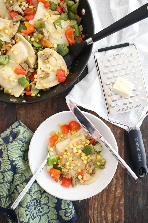 pierogies with onions, peppers, and sausage in a white bowl and a black pan, with cheese and a grater