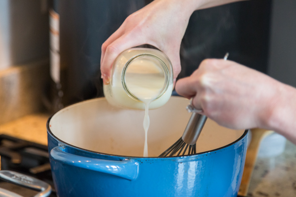 milk being poured into a blue dutch oven to make baked pasta with sausage and broccolini
