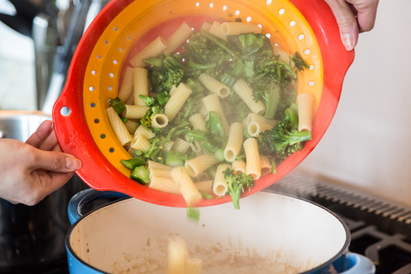 broccoli and pasta being poured from a colander to a blue dutch oven to make baked pasta with sausage and broccolini