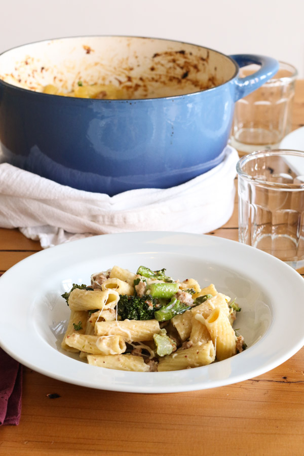 baked pasta with sausage and broccolini in a white bowl set on the dinner table with a blue dutch oven