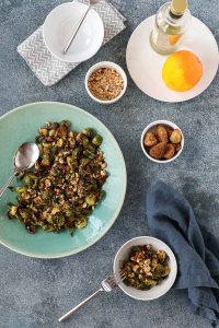 roasted brussels sprouts with bacon and figs on a teal plate with small bowls of figs, hazelnuts, and an orange