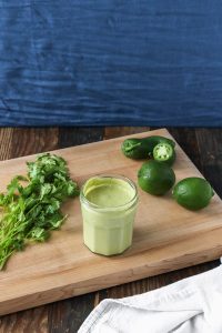 creamy jalapeño cilantro sauce in a glass jar with cilantro, jalapeños, and limes on a wooden board