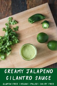 creamy jalapeño cilantro sauce in a glass jar with cilantro, jalapeños, and limes on a wooden board