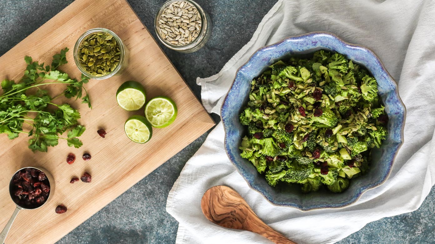 spicy miso tahini broccoli salad with dried cranberries, cilantro, limes, sunflower seeds, and pepitas on a wooden cutting board